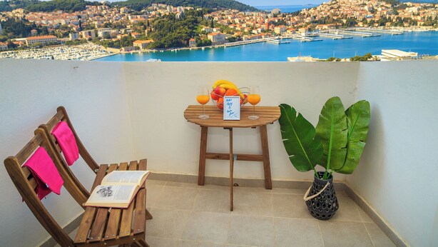 A balcony with a wooden table, a chair, a potted plant, and a view of a coastal town.
