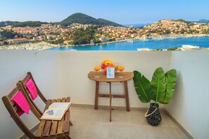 A balcony with a wooden table, a chair, a potted plant, and a view of a coastal town. - Skyscraper Apartment- Free parking included (Dubrovnik)