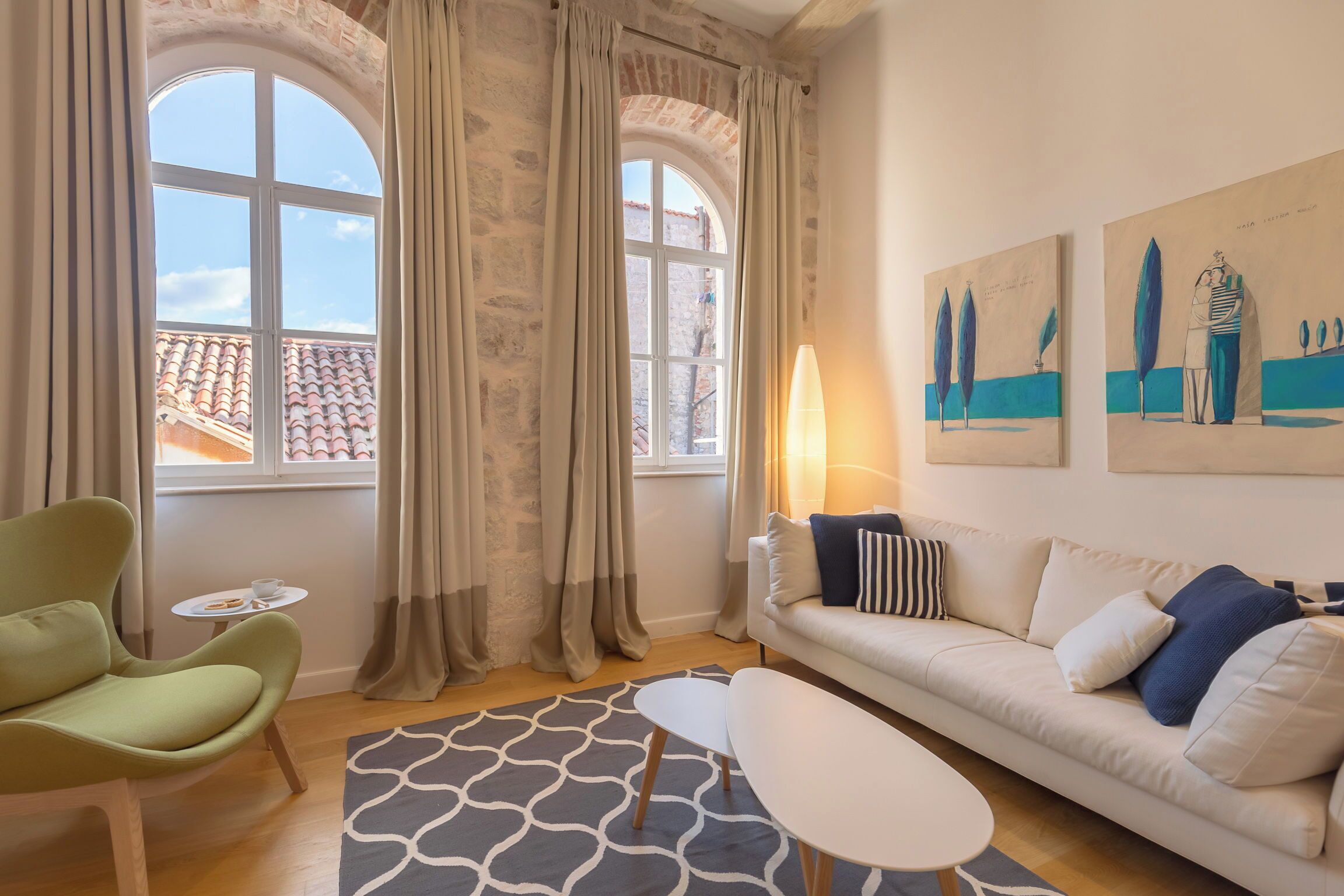 Een moderne woonkamer met een groene fauteuil, een witte bank en een kamerkleed met een patroon. Er zijn twee ramen met gordijnen, een klein tafeltje met een kopje en een lamp.