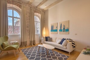Un salon moderne avec un canapé, un fauteuil et une table basse. Il y a deux fenêtres avec des rideaux, une lampe et des œuvres d’art accrochées au mur.