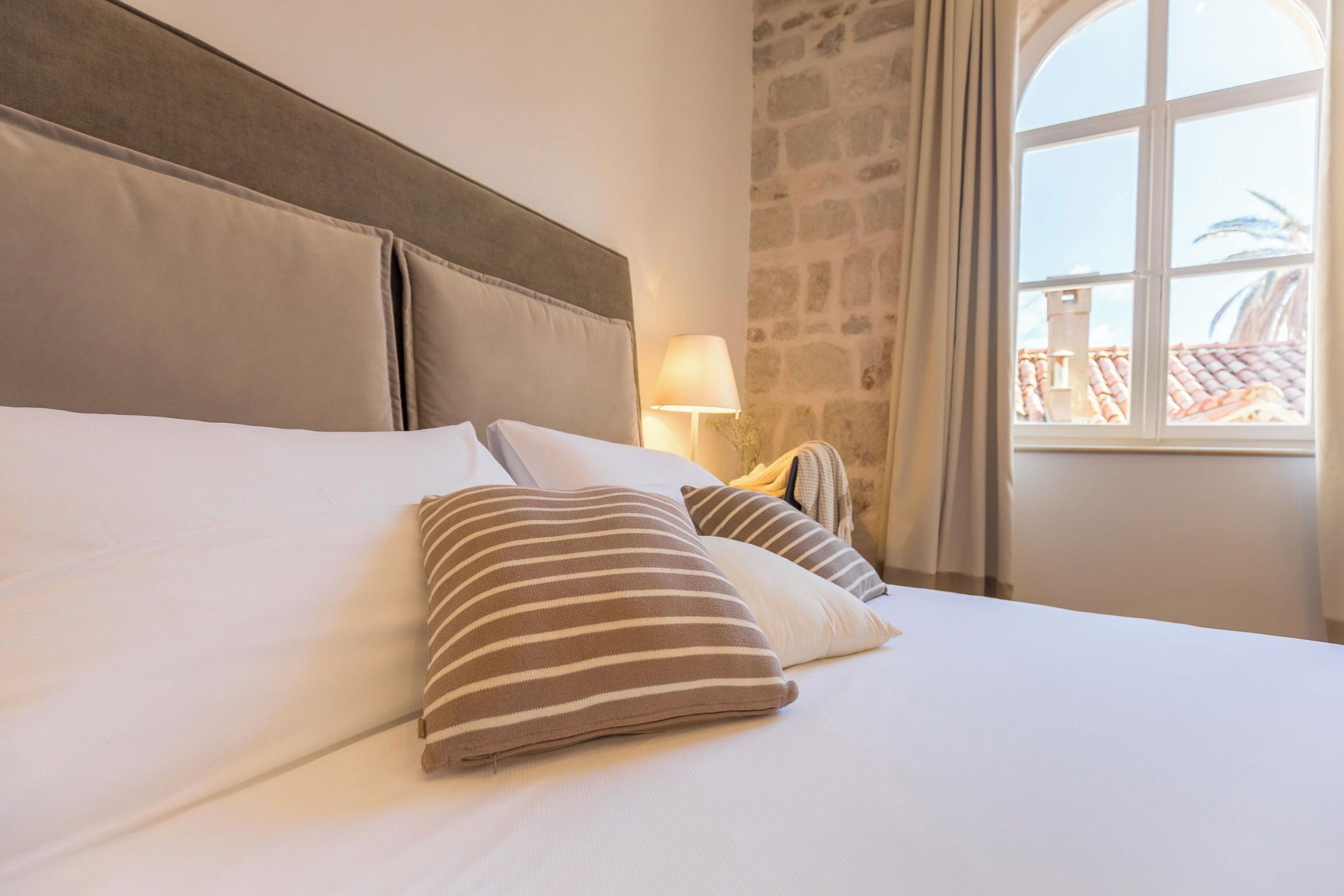 Ett hotellrum med en stor säng, en vägg av sten och ett fönster med utsikt över byggnader.