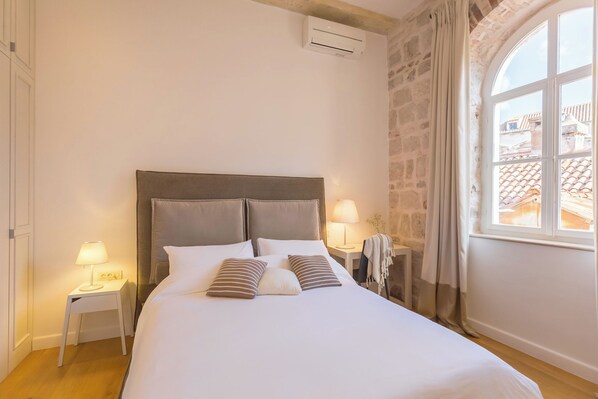 Una cama bien tendida con un cabecero, dos mesitas de noche con lámparas, una ventana con cortinas y una pared de piedra.
