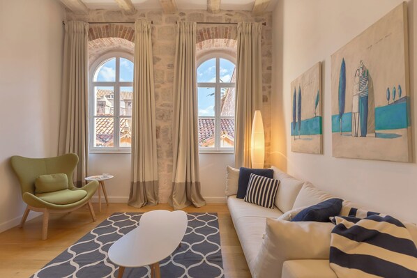 Un salon chaleureux avec un fauteuil vert, un canapé blanc et un tapis à motifs. Il y a deux fenêtres avec des rideaux, une petite table et une lampe.