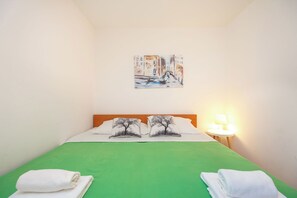 A hotel room with a green bedspread, two pillows with tree designs, a painting of a canal scene, and a bedside lamp.
