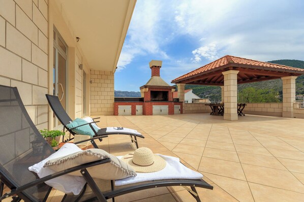 A patio with two lounge chairs, a sun umbrella, a grill, and a view of a mountain.