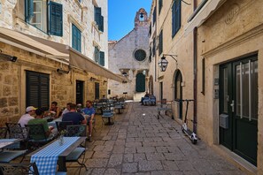 Una calle estrecha con mesas al aire libre, edificios de piedra y una torre de campanas.