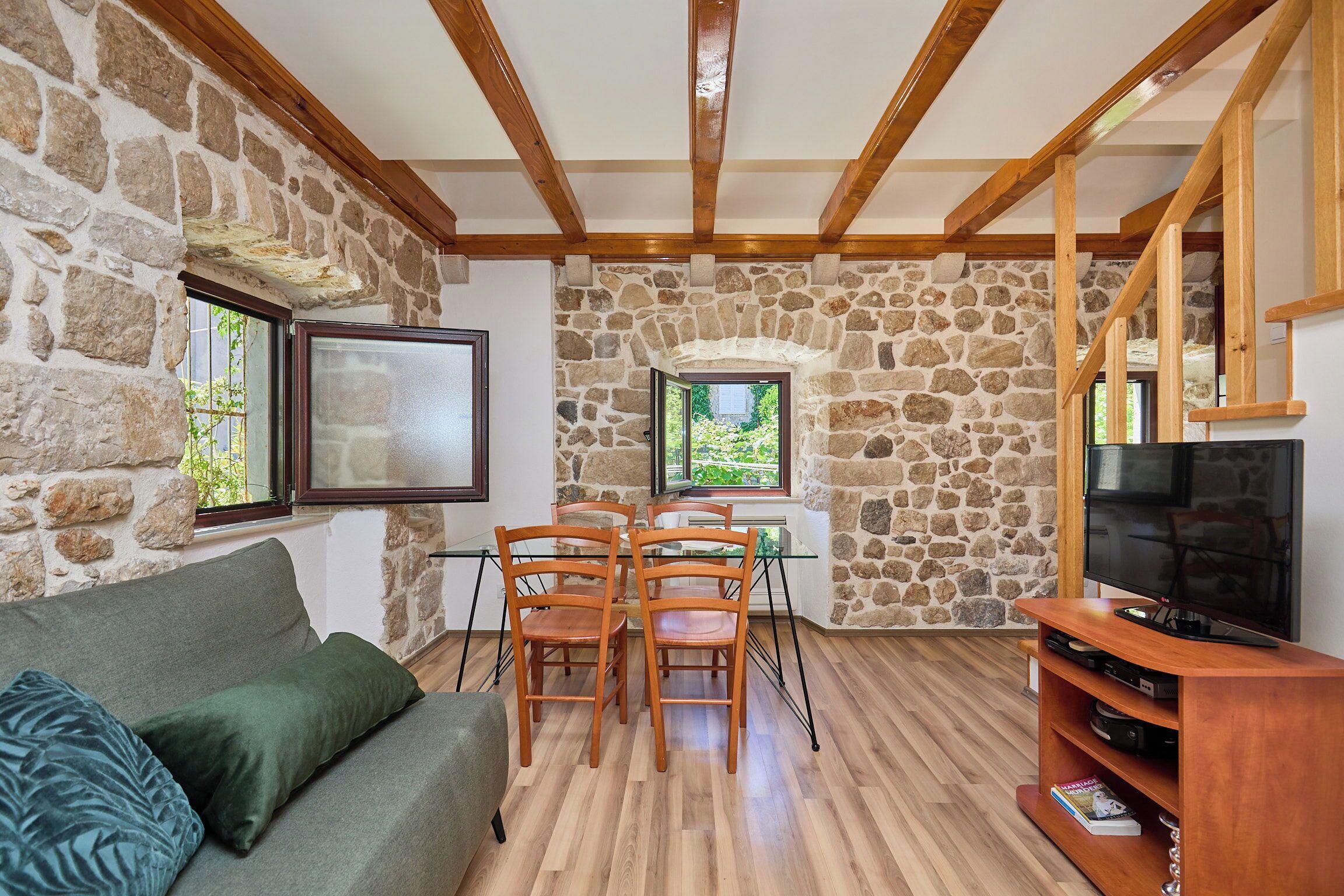 Una sala de estar acogedora con pared de piedra, vigas de madera, una mesa de vidrio con sillas, un mueble para televisión y un sofá con cojines.