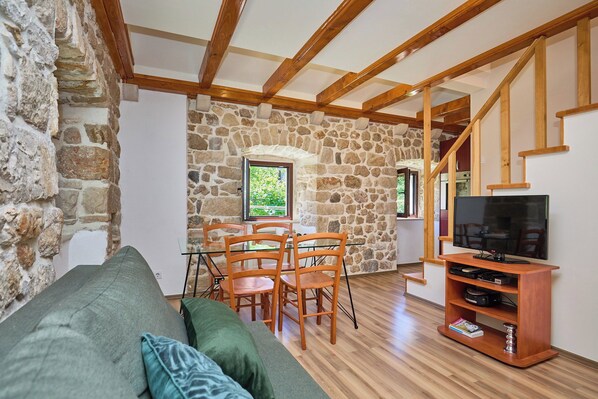 Una sala de estar acogedora con pared de piedra, vigas de madera en el techo, un sofá verde, una mesa de comedor con sillas y un mueble para televisión.