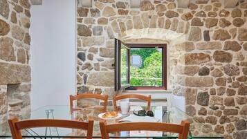 Una habitación con paredes de piedra, una mesa de vidrio y sillas, una ventana con vistas a la vegetación y una puerta.