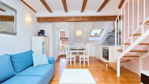 Un salon moderne avec un canapé bleu, une table basse blanche et un coin repas comprenant une table et des chaises. On y trouve également une cuisine équipée d’une cuisinière et un escalier blanc.
