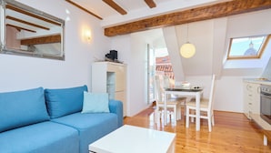 Un salon avec un canapé bleu, une table basse blanche, un coin repas avec une table et des chaises, une cuisine équipée d’un four et une lucarne offrant une vue sur un immeuble.