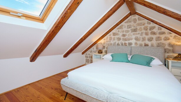 Une chambre avec un plafond en pente, un mur en pierre, des poutres en bois, un lit avec une literie blanche et une table de chevet avec une lampe.