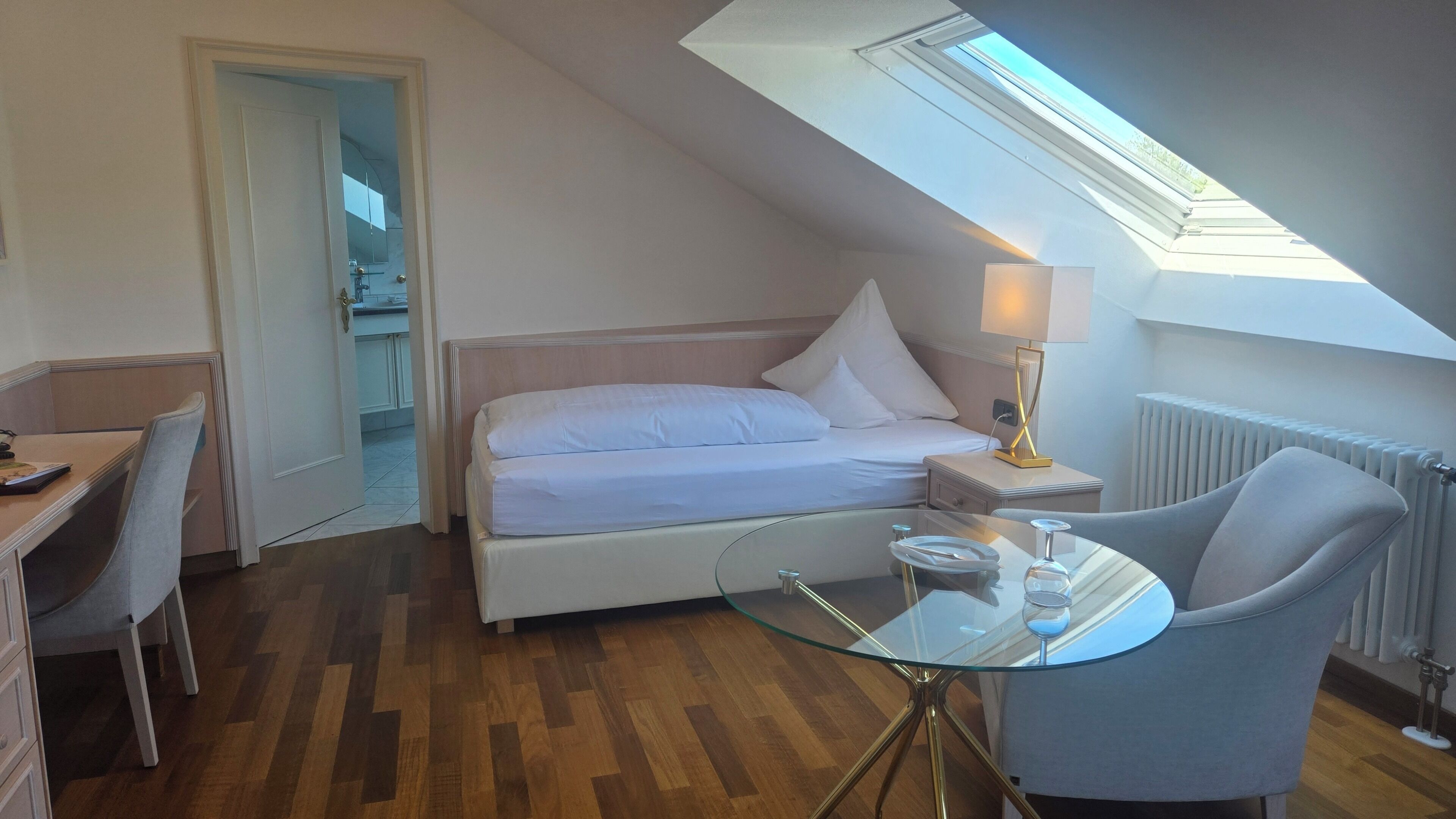 A bedroom with a bed, a glass table, a chair, a lamp, and a skylight.