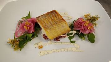 A white plate with a piece of food, possibly a pastry, surrounded by greens and pink flowers.