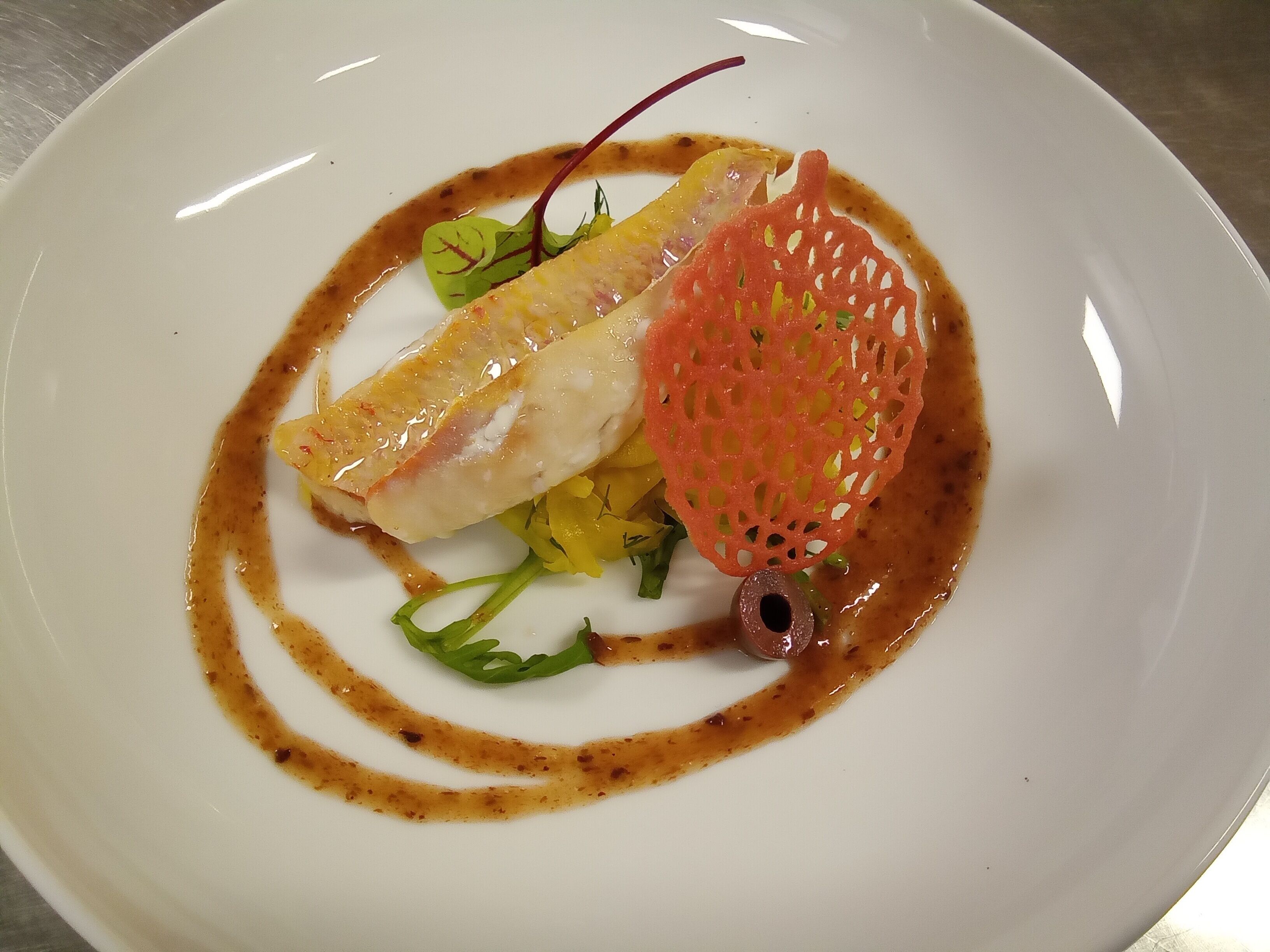 Ein weißer Teller mit Fischfilet, grünem Gemüse und einem roten dekorativen Element.