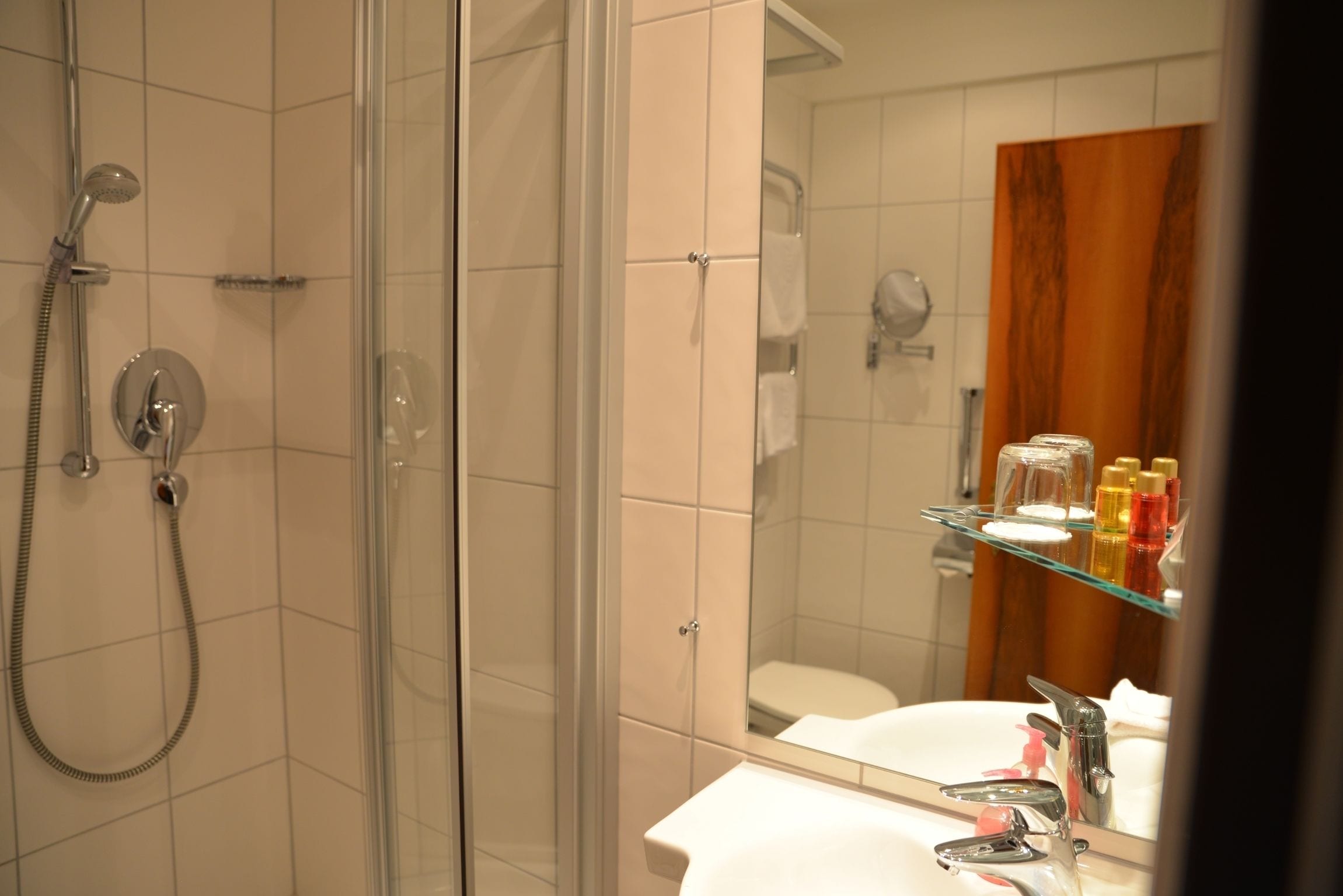 Un baño con ducha de vidrio, lavamanos blanco con espejo y repisa con artículos de aseo.