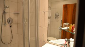 Un baño con ducha de vidrio, lavamanos blanco con espejo y repisa con artículos de aseo.