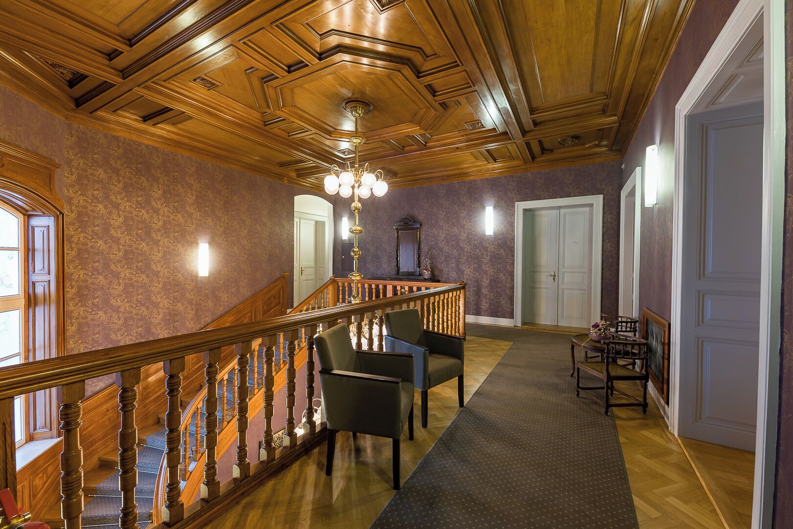 Un amplio pasillo con una escalera de madera, un candelabro y una sala de estar con sillones y una mesa.