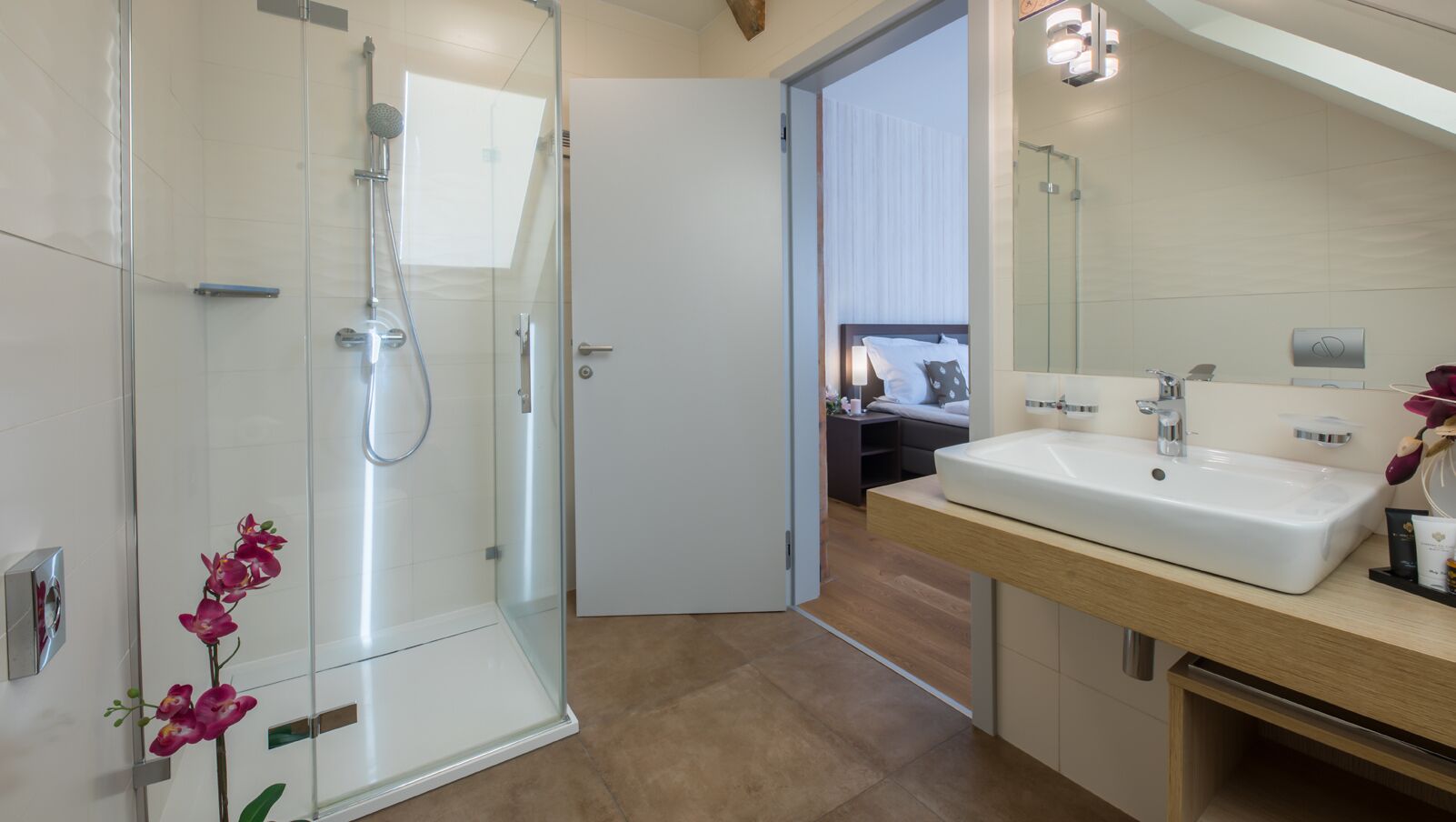 A modern bathroom with a glass shower enclosure, a wooden vanity with a sink, and a visible bedroom in the background.