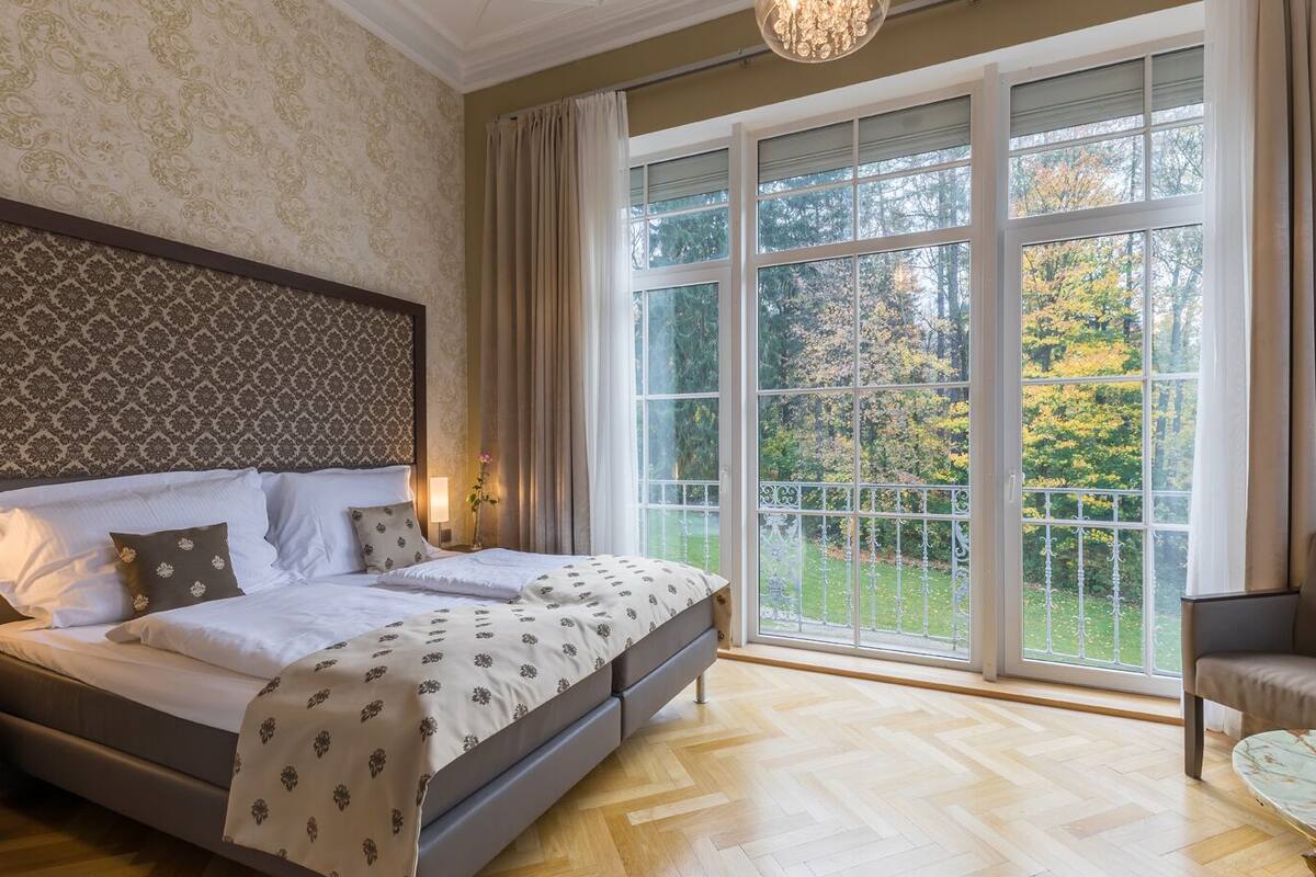 Ein Schlafzimmer mit einem großen Bett, einem Sessel und Blick auf einen Garten durch große Fenster.