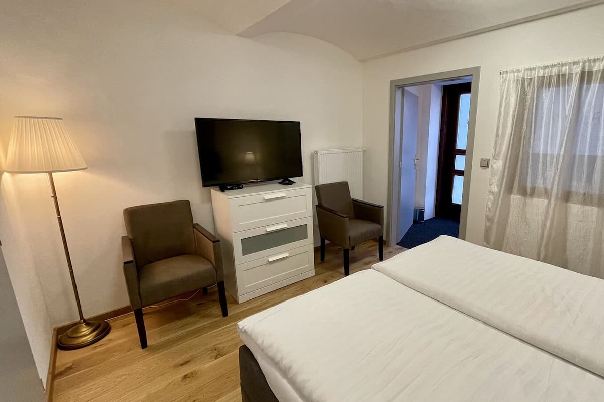 Ein Hotelzimmer mit einem Bett, einem Fernseher auf einer Kommode, zwei Sesseln, einer Stehlampe und einer Tür zu einem weiteren Raum.