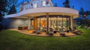 Uma casa moderna com uma extensão de vidro curvo, cadeiras de lounge ao ar livre e um gramado bem cuidado.