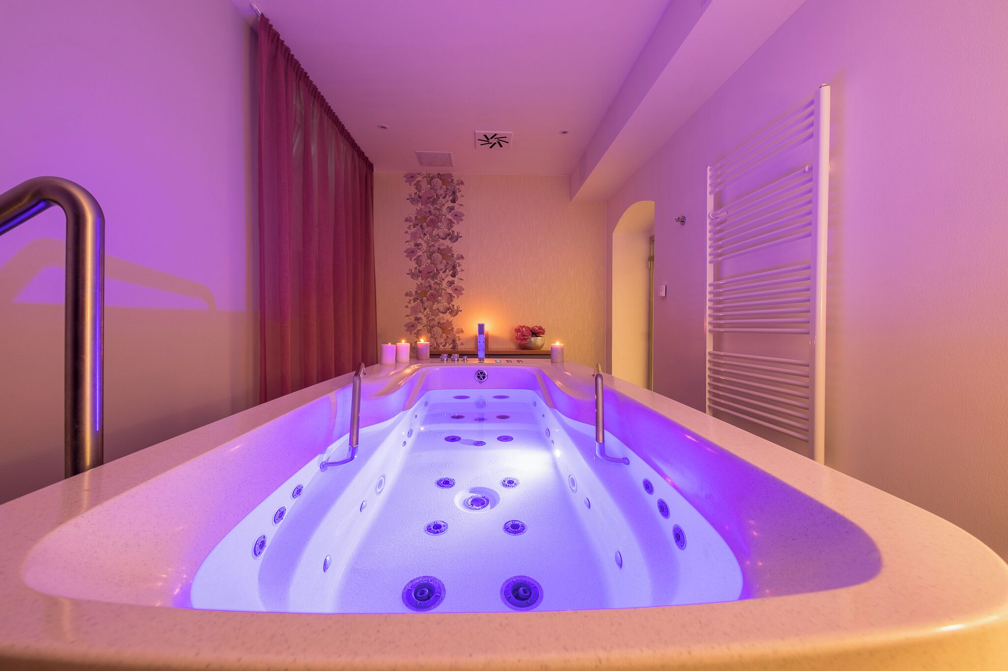 Een bubbelbad met een paars verlicht interieur, een wandheater en een klein tafeltje met kaarsen.