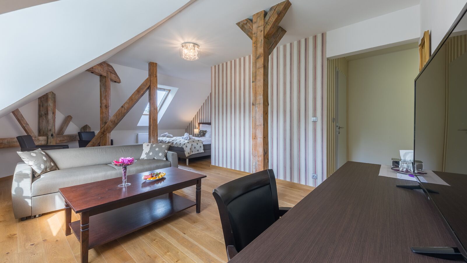 Een moderne loft slaapkamer met een schuine plafond, houten balken, een groot bed, een zithoek met een bank en salontafel, en een eettafel.