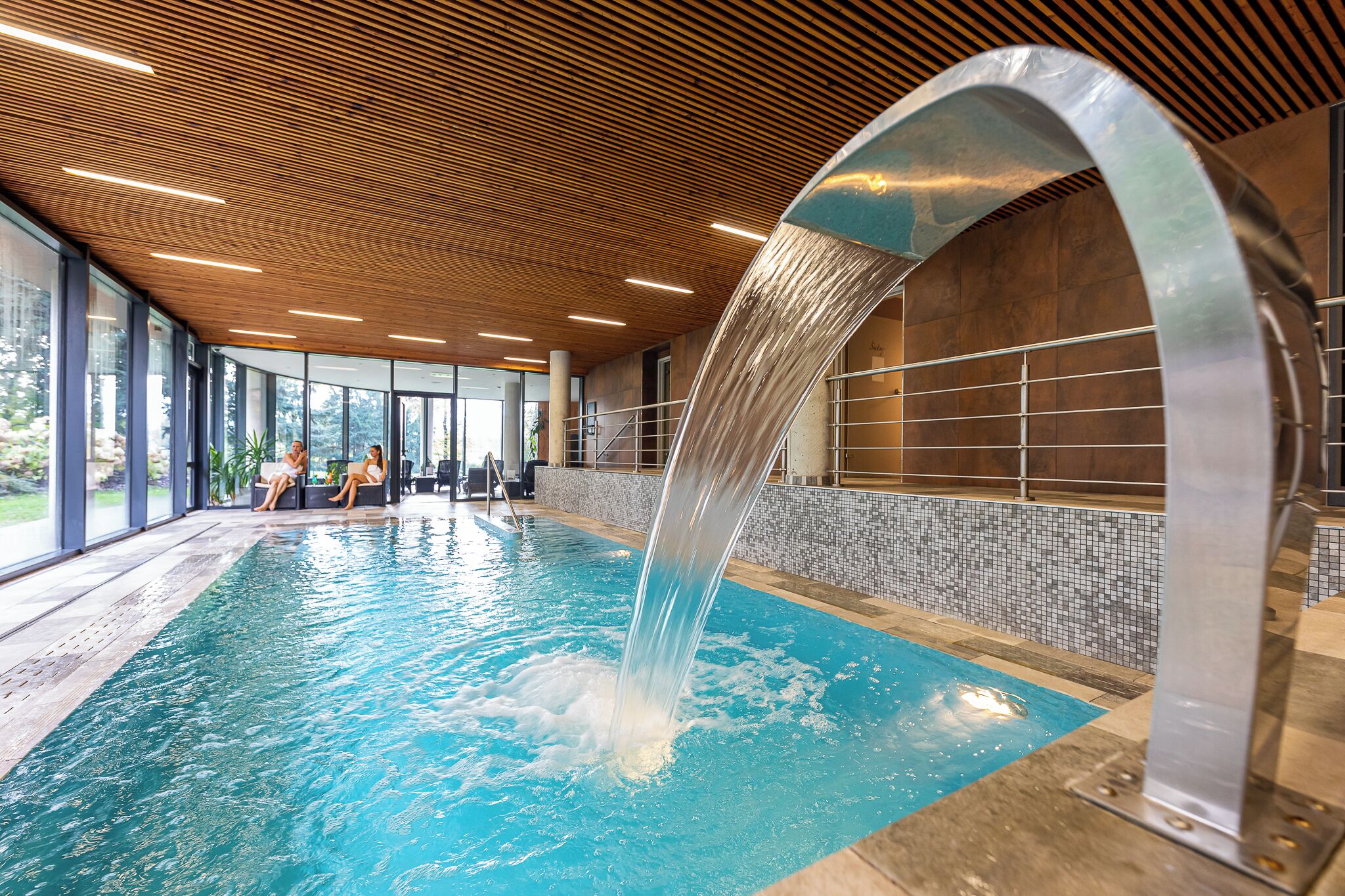 An indoor swimming pool with a water feature, large windows, and a wooden ceiling.