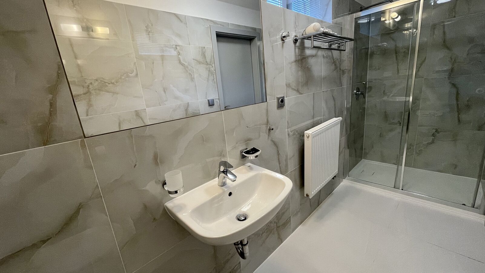 Une salle de bain moderne avec un mur recouvert de carreaux de marbre, un lavabo blanc, une douche en verre et une petite fenêtre.