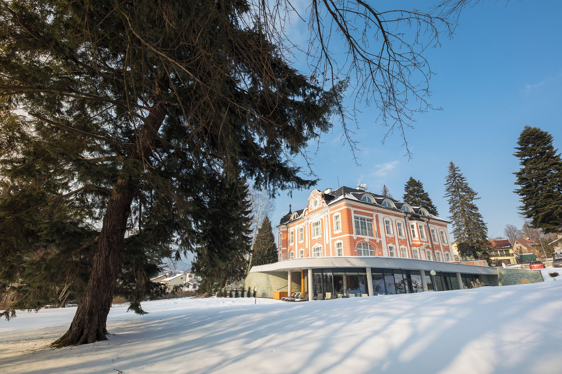 Un edificio grande e histórico con un ala de vidrio curvo, rodeado de nieve y altos árboles de hoja perenne.