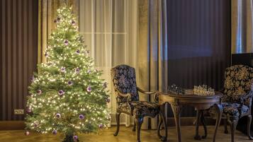 A Christmas tree with lights and ornaments, a chess table, and two upholstered chairs in a room with wooden flooring and curtains.