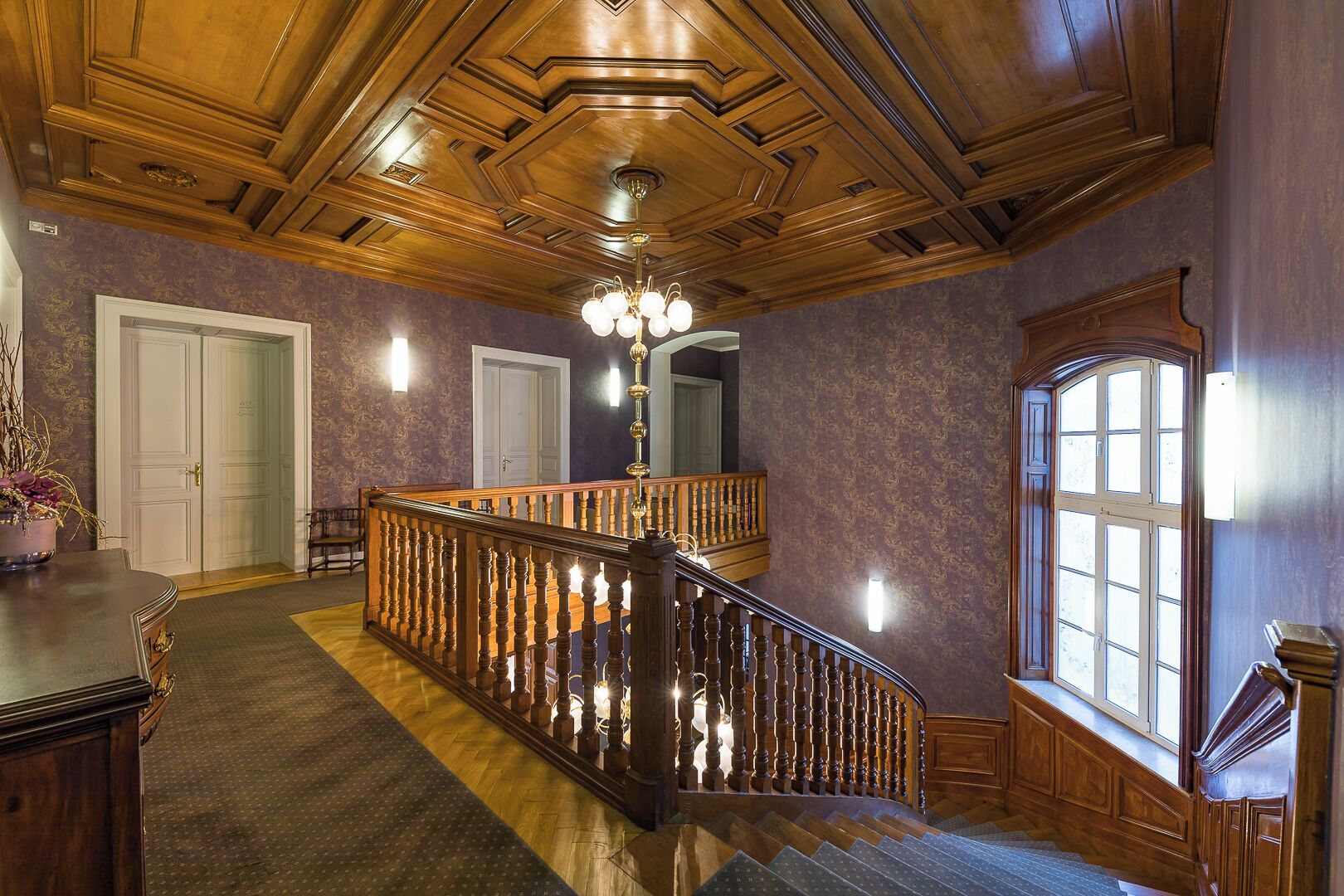 A grand staircase with a wooden railing, a chandelier, and a large window with multiple panes.