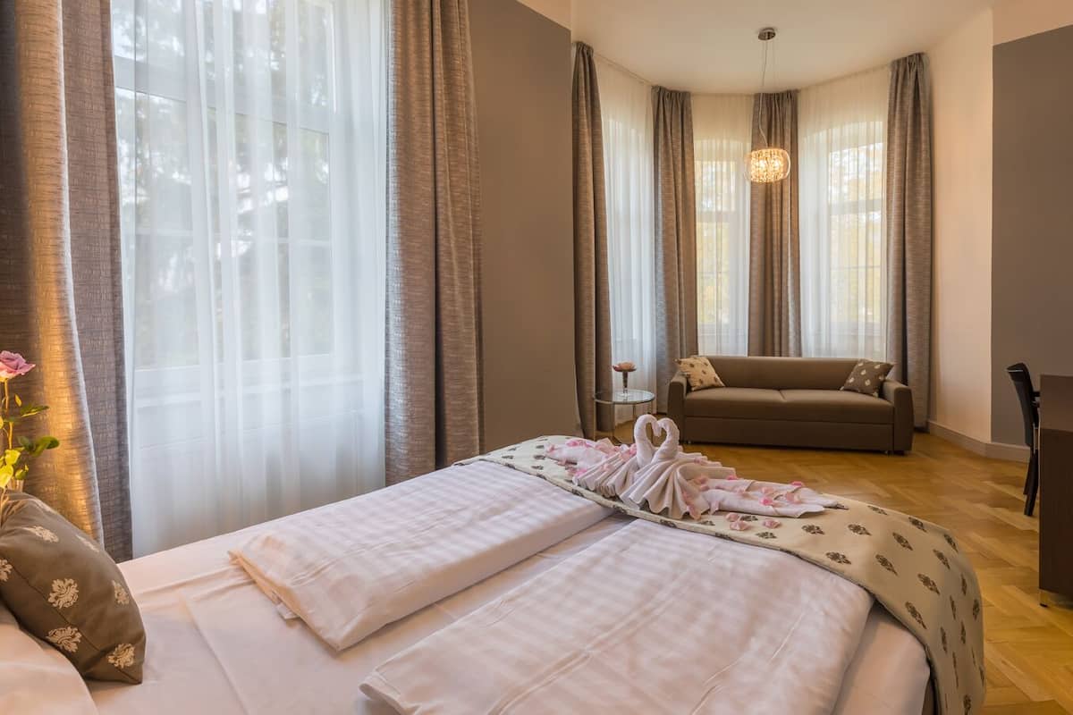Ein Hotelzimmer mit zwei Betten, einem Sofa, einem Schreibtisch und einem großen Fenster mit Vorhängen.