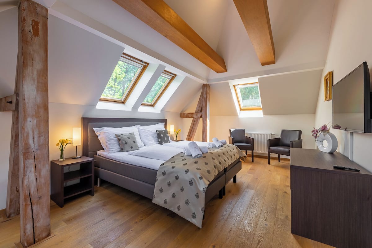 Ein Schlafzimmer mit Bett, Nachttischen, Schreibtisch, Stuhl und Fernseher. Im Deckenbereich befinden sich Dachfenster und Holzbalken.