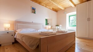 Une chambre avec un lit en bois, des tables de chevet blanches, une lampe de chevet, une porte verte et une fenêtre donnant sur un paysage verdoyant.