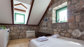 Une chambre avec un mur en pierre, des poutres en bois, un lit avec du linge de lit blanc, une armoire blanche et une fenêtre avec un cadre vert.