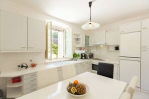 Une cuisine moderne avec des meubles blancs, une table à manger agrémentée d’un panier de fruits et une fenêtre donnant sur un paysage verdoyant.
