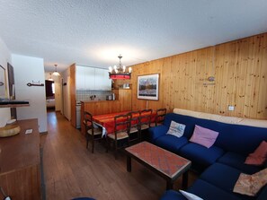 Een gezellige woonkamer met een blauwe bank, een houten eettafel en stoelen. Achtergrond toont een keuken met witte kastjes en een hangende lamp.
