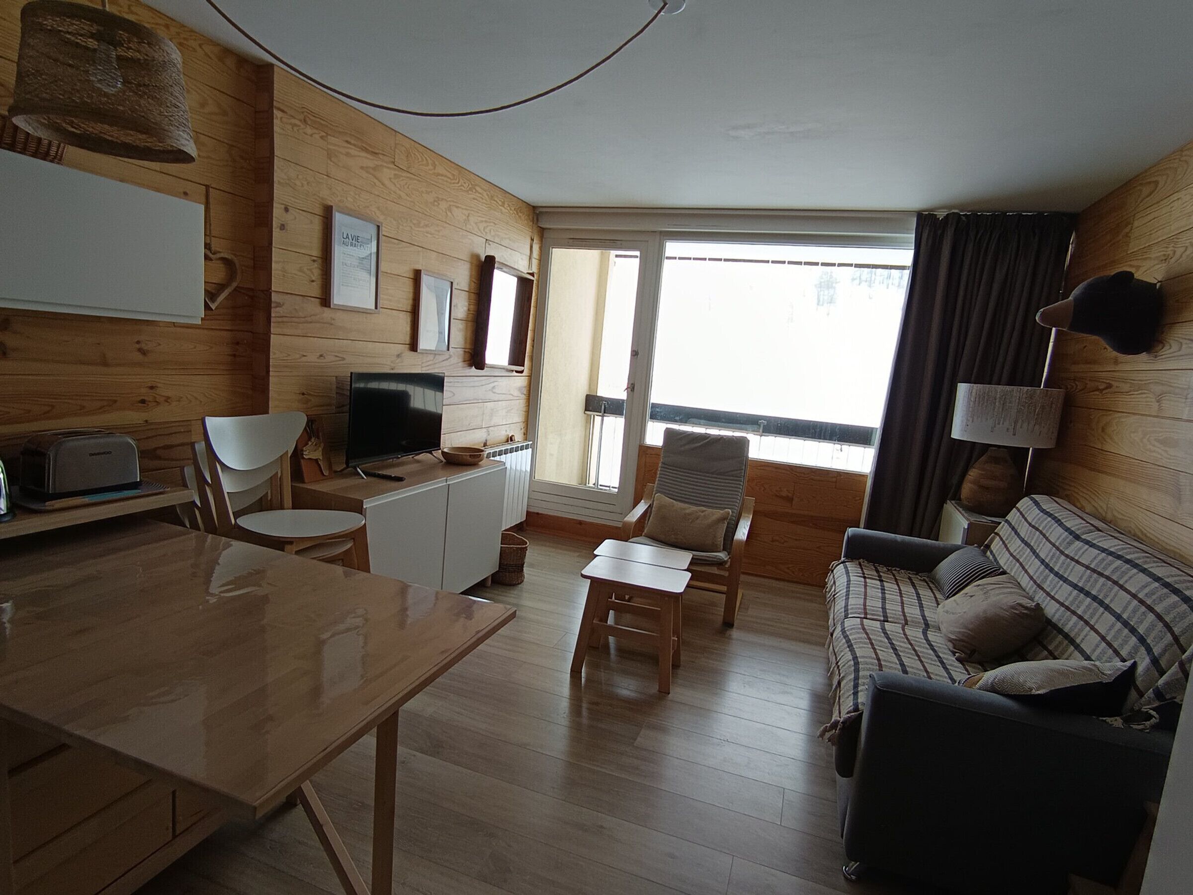 A cozy wooden interior with a dining table, a TV, a sofa, and a balcony with a view.