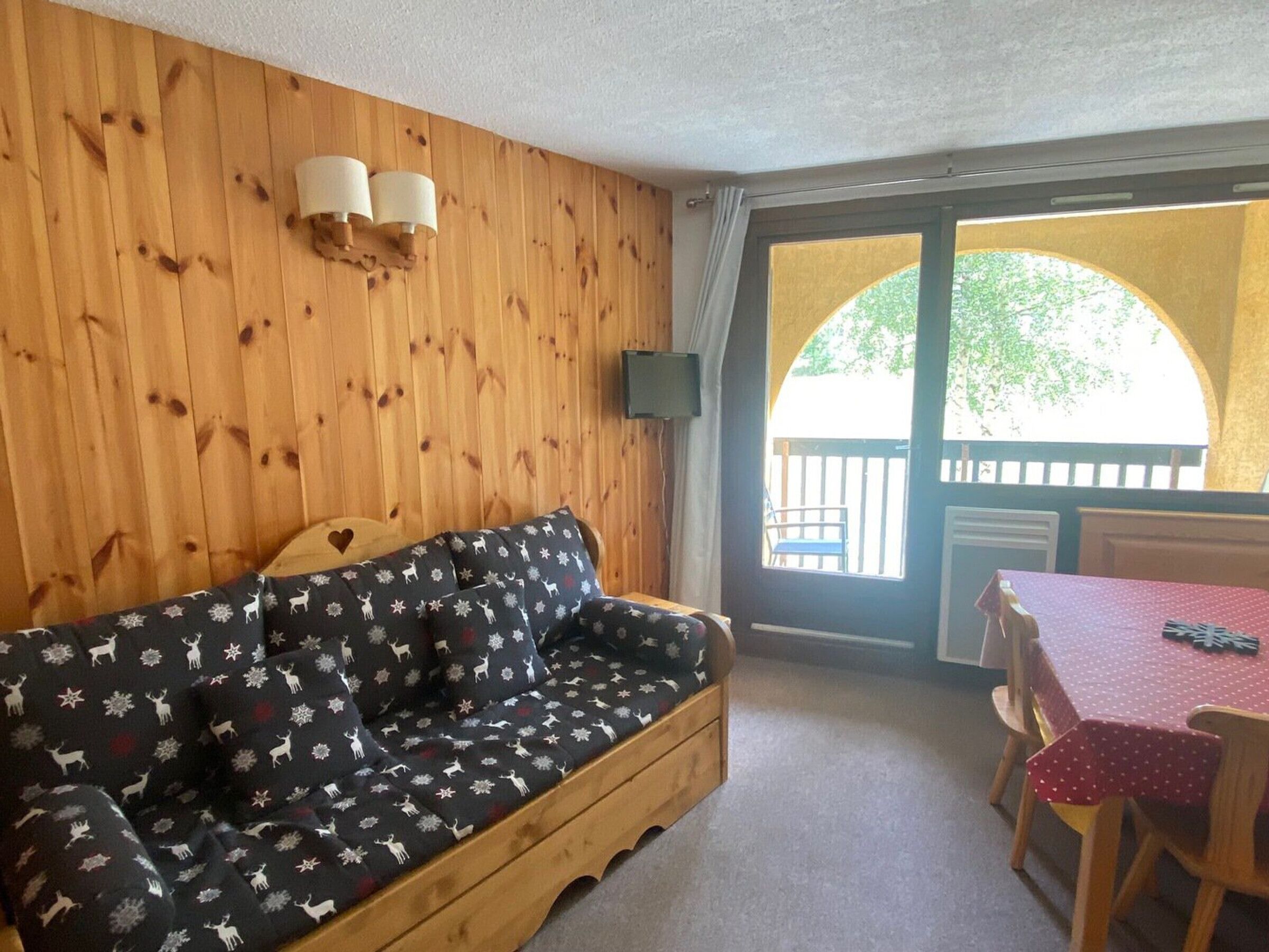 A cozy room with a wooden wall, a sofa with a festive pattern, a table with a red tablecloth, and a view of an outdoor deck.