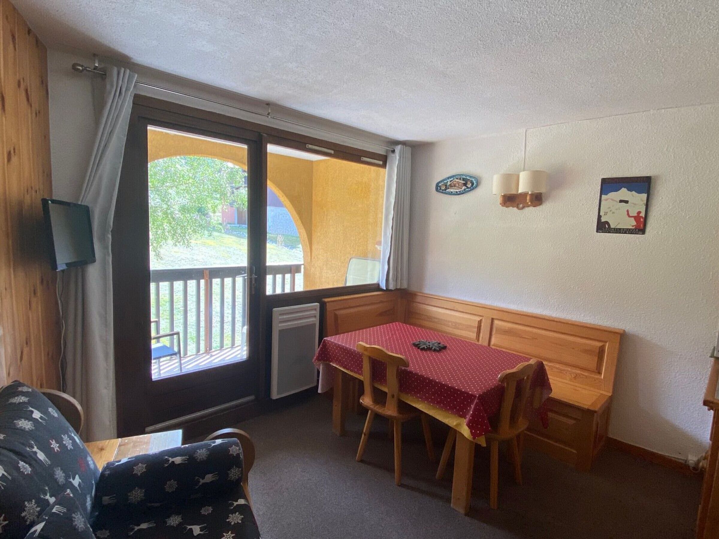 A small room with a wooden bench and table, a sliding glass door to a balcony, and a small TV mounted on the wall.