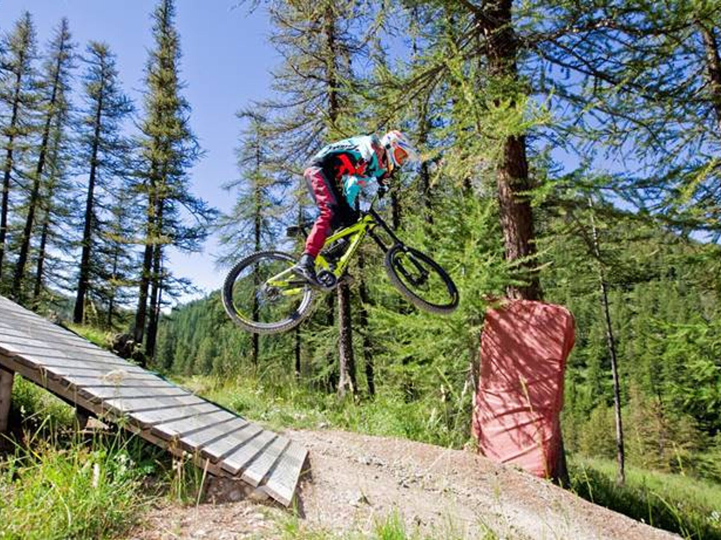 A mountain biker performing a jump over a wooden ramp in a forested area.