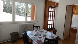 Un coin repas avec une table recouverte d’une nappe aux motifs de café, deux chaises, un radiateur, une fenêtre donnant sur des arbres et une porte en bois.