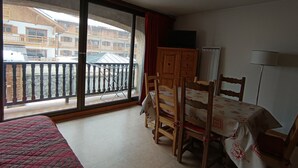 A room with a bed, a table with chairs, a wooden cabinet, and a large window with a balcony view.