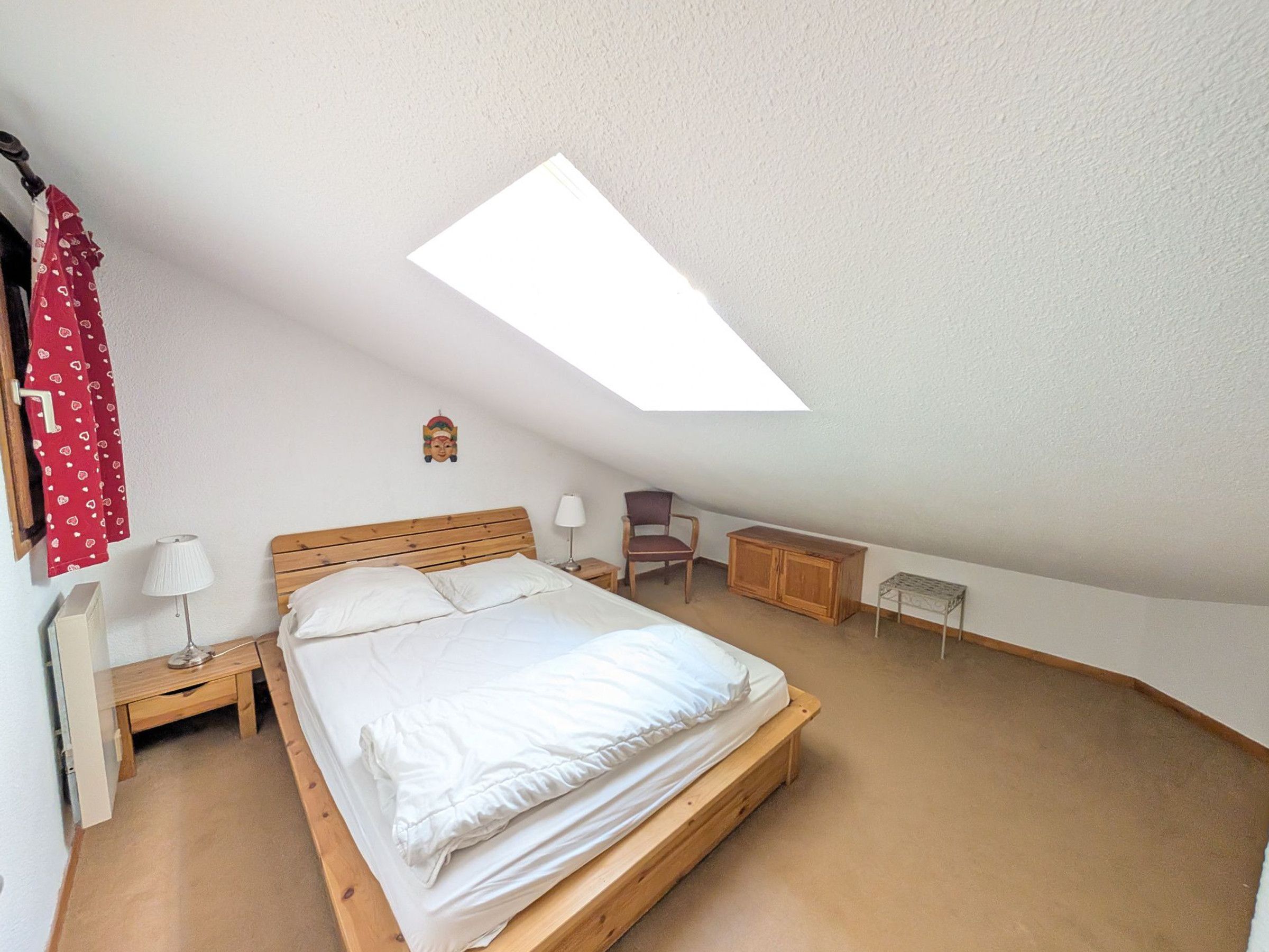Une chambre avec un plafond en pente, un lit, une table de chevet, une chaise, une petite table et une armoire.