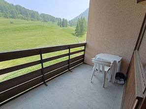 A balcony with a view of a green field and mountains. There is a small table and a chair on the balcony.