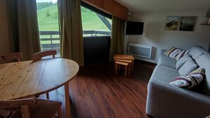 A room with a wooden table and chairs, a sofa, a television, and a window with curtains. - Lovely 2P 5 Pers Ski-in Ski-out Top Floor Pets Welcome (Montgenèvre)