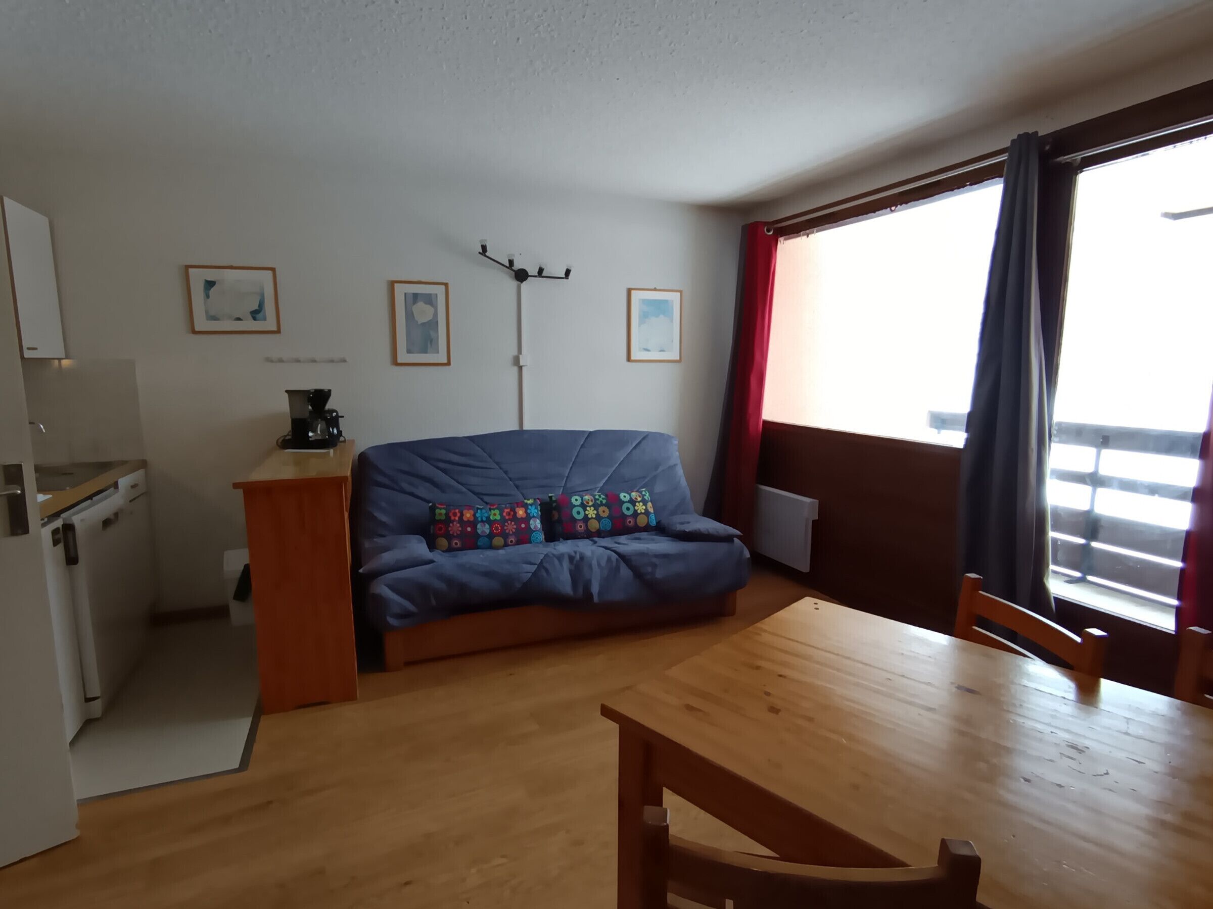 A compact living space with a kitchenette, a sofa with colorful cushions, a dining table, and a large window with curtains.