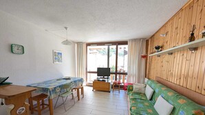 A cozy living room with a wooden sofa, a dining table with chairs, a TV, and a large window with curtains. - Studio with south-facing balcony, parking in Montgenèvre, pets allowed (Montgenèvre)
